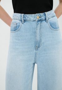Jeans de denim azul claro de talle alto con un botón frontal, presillas para cinturón y dos bolsillos laterales. Presenta una textura limpia y suave.