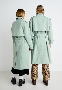 Duas pessoas vestem casacos trench verde menta com cintos na cintura, apresentando um design de duplo botão e abas nos ombros, combinados com calças estampadas.