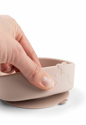 Hånd griber en beige silikoneskål med prægede kløverformer og en sugekopbase på en hvid overflade.