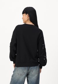 Black sweatshirt made of soft fabric, featuring cut-out floral designs on both sleeves and a relaxed fit. Paired with light blue jeans.