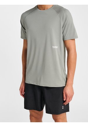 Mann trägt hellgraues Sportshirt mit kurzen Ärmeln und schwarze Shorts vor weißem Hintergrund, steht mit entspannten Armen seitlich am Körper.