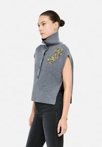 Maglione grigio a maglia con colletto alto, maniche corte e applicazione decorativa in foglia dorata. Presenta chiusura con bottoni e texture a coste.