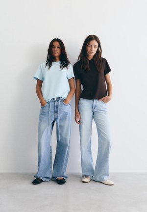 Două femei stau sprijinite de un zid alb, purtând blugi largi, de culoare albastru deschis; una poartă un tricou albastru deschis și pantofi negri, cealaltă un tricou negru și pantofi albi.