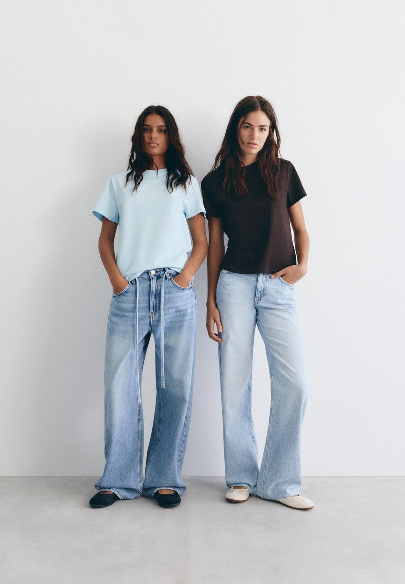 Deux femmes se tiennent contre un mur blanc, portant des jeans larges bleu clair, l'une avec un t-shirt bleu clair et des chaussures noires, l'autre avec un t-shirt noir et des chaussures blanches.