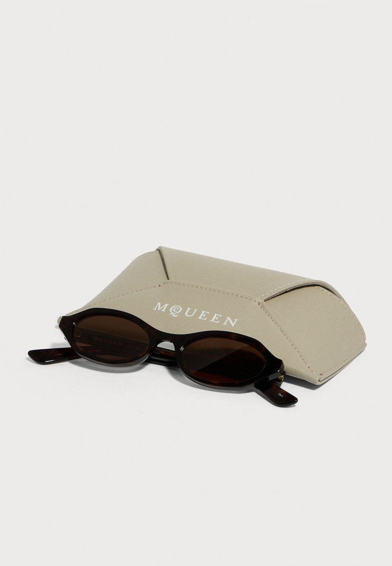 Ovale Sonnenbrille mit dunklen Gläsern, liegend vor einem beigen, mit MQUEEN beschrifteten Schutzetui auf weißem Hintergrund.