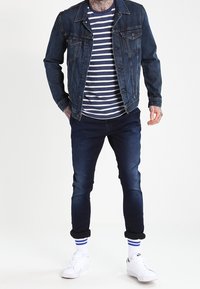 Jeansjacka över en marinblå och vit randig skjorta, kombinerad med mörka jeans. Vita sneakers med blårandiga strumpor.