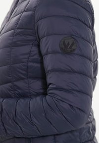 Marinblå pufferjacka med kviltad textur, försedd med en rund svart logopatch märkt "WHISTLER." Sittande design med hög krage.