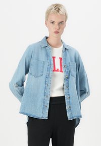 Camisa de denim azul claro com colarinho, fecho de botões à frente, dois bolsos no peito, mangas arregaçadas e um corte descontraído. Usada sobre uma T-shirt branca.