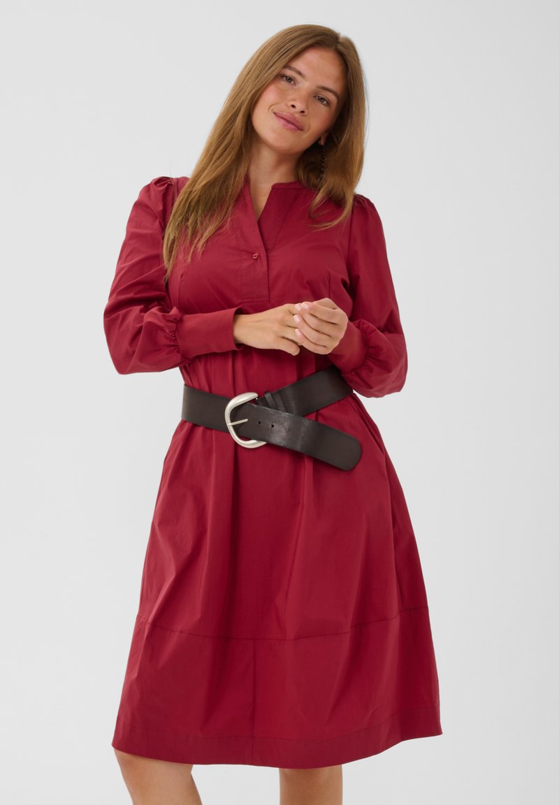 Femme aux longs cheveux châtain clair portant une robe rouge à manches longues et une large ceinture noire, debout, mains jointes.