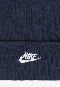 Tamno plavi pleteni beanie izrađen od mekog materijala, s bijelim izvezenim Nike logotipom sprijeda i dizajnom s preokrenutim rubom.