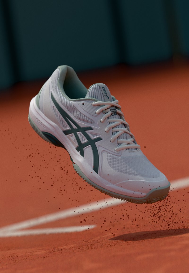 White athletic shoe with mesh detailing, grey accents, and a textured sole, suspended above a red surface, with dirt particles dispersing.