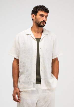 Man met kort donker haar en baard, draagt een wit overhemd met korte mouwen en abstracte borduursels over een olijfgroene tanktop.