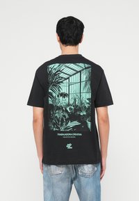 Άνδρας με μαύρο T-shirt με γαλάζιο-πράσινο γραφικό ενός θερμοκηπίου γεμάτου φυτά, σε συνδυασμό με ανοιχτό μπλε ξεβαμμένα τζιν, στέκεται μπροστά σε λευκό φόντο.