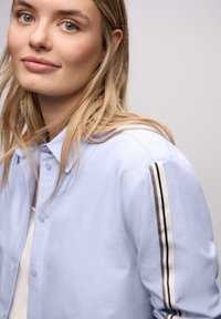 Lyseblå skjorte med knapper, der har en klassisk krave, blødt stof og stribede detaljer i brunt og hvidt på ærmerne.