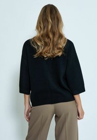 Pull en tricot noir avec une coupe ample et des manches trois-quarts, associé à un pantalon beige à jambes larges. Le tissu est doux avec une texture subtile.