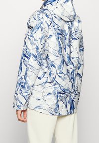 Chaqueta resistente al agua en blanco con un patrón floral azul; cuenta con capucha, cierre con cremallera y puños elásticos. Textura suave con un diseño para el aire libre.