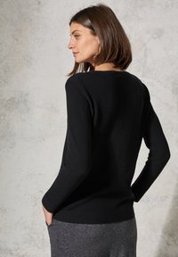 Schwarzer, gerippter Pullover mit Rundhalsausschnitt und langen Ärmeln, der ein enganliegendes Design und einen strukturierten Stoff aufweist. Der Hintergrund ist neutral und strukturiert.