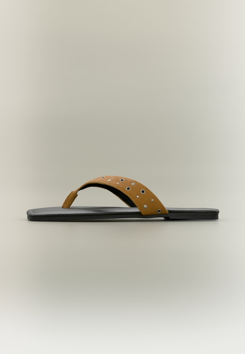 Φλατ μαύρο σανδάλι με καφέ δερμάτινο λουράκι στα δάχτυλα, διακοσμημένο με ασημένιες τρουκς και μεταλλικά ρουμπάκια σε απλό φόντο.