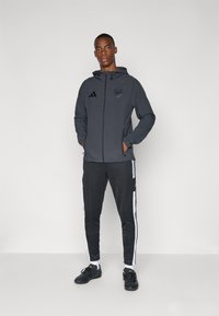 Svart atletisk jakke med hette og glidelås, prydet med Arsenal-logoen og Adidas-merket. Kombinert med svarte treningsbukser og svarte sko.