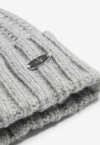 Šedá pletená čepice má ribbovaný povrch, ohnutý lem a stříbrný oválný štítek připevněný bílou nití.