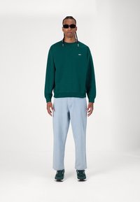 Green sweatshirt with logo, light blue loose-fit jeans, and dark sneakers. The model wears black sunglasses and has braided hair.