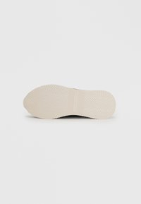 Black sneaker sole with herringbone traction pattern, smooth texture, and a cream-colored rubber bottom. Features logo detail in the center.