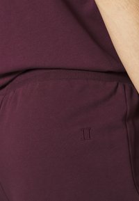 Gros plan d'une personne portant un pantalon et un haut de couleur violet foncé avec une taille élastique et un petit logo brodé "II" sur le pantalon.
