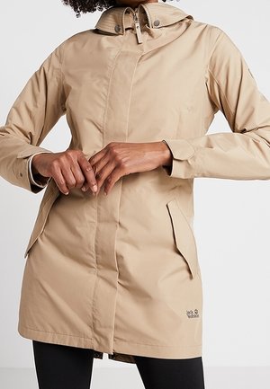 Beżowy płaszcz przeciwdeszczowy z kapturem, zapinany na zamek i zatrzaski, z przednimi kieszeniami na klapę, noszony przez osobę ze splecionymi rękami na wysokości talii.