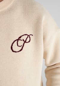 Pulover bej tricotat cu un guler înalt, prezentând un logo brodat în roșu închis, format din literele "F" și "P" intercalate, pe piept.