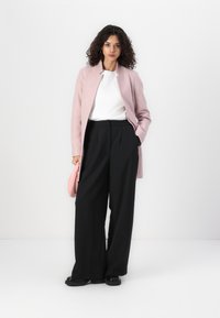 Cappotto su misura rosa chiaro sopra una maglietta bianca, abbinato a pantaloni neri a gamba larga con vita alta. Tiene una piccola borsa a mano rosa.