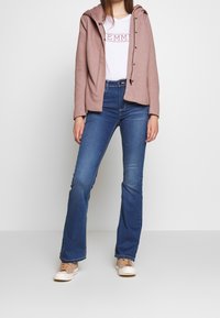 Lichtroze gewatteerde jas met een rits aan de voorkant, witte t-shirt met de tekst "EMMA", blauwe flared jeans en beige sneakers.