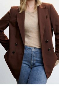 Personne portant un blazer marron croisé sur un pull beige côtelé et un jean bleu taille haute, mains dans les poches du blazer.