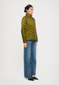 Kvinne står med siden til, iført en olivenfarget quiltet jakke, vide blå jeans og svarte spisse lavhælte sko.