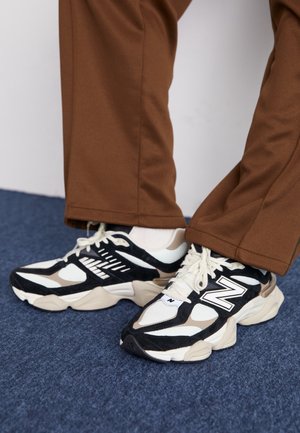Osoba nosząca czarne, białe i beżowe sneakersy New Balance w połączeniu z brązowymi spodniami, stojąca na niebieskim dywanowym podłożu.