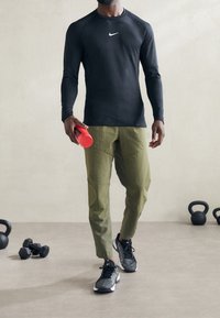 Černé sportovní tričko s dlouhým rukávem, olivově zelené zúžené kalhoty, černo-bílé běžecké boty, drží červenou láhev na vodu, za ním činky.