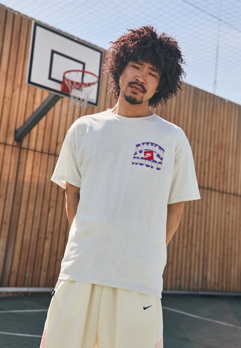 Lys grå bomulds t-shirt med et multicolor "Nike Hoops" grafisk design på brystet, parret med creme shorts med et sort logo.