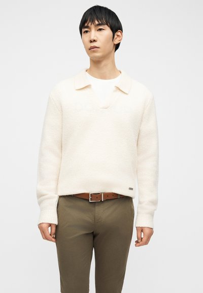 Άνδρας με εκρού πουλόβερ πάνω από λευκό πουκάμισο, καφέ ζώνη και ελιά παντελόνι, στέκεται μπροστά σε απλό λευκό φόντο.