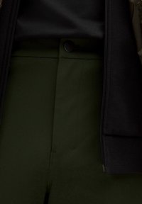 Tumed rohelised püksid on lamedate esiosadega, musta nööbiga kinnitatud. Kangal on sile välimus kerge läikega, mida on sobitatud musta ülikonnaga.