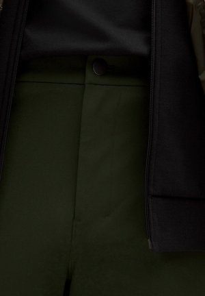 A sötétzöld nadrág sima elülső kialakítással és fekete gombzárral rendelkezik. Az anyaga simának tűnik, egy kis fényt is mutat, fekete felsővel párosítva.