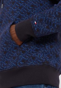 Felpa blu navy con un intricato motivo blu, dotata di tasca a marsupio, polsini a coste e un piccolo logo rosso, bianco e blu sulla manica.
