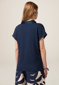 Tricou bleu cu mânecă scurtă, cu guler din satin și hem rotunjit. Țesătură netedă cu o textură subtilă, combinată cu pantaloni cu un model ce prezintă frunze mari.
