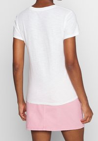 Bílá tričko s krátkým rukávem a kulatým výstřihem, s mírně strukturovanou tkaninou. V kombinaci se světle růžovou džínovou sukní.
