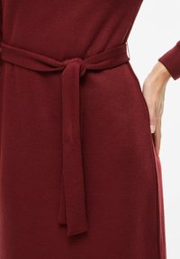 Bordowa sukienka prążkowana z przewiązywanym paskiem w talii i kieszeniami po bokach, wykonana z fakturowanej tkaniny o dopasowanym fasonie.