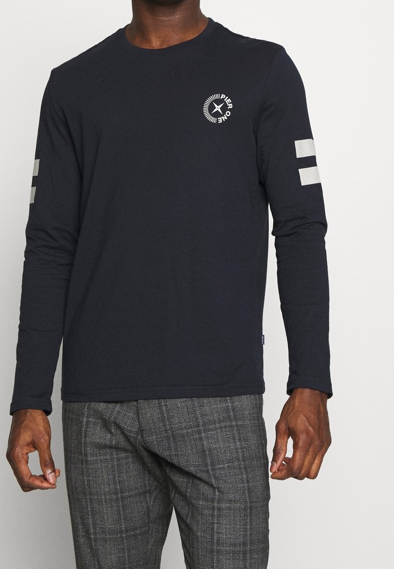 Man i en långärmad marinblå skjorta med "Pier One" cirkellogo på bröstet och vita ränder på ärmarna, tillsammans med grå rutiga byxor.