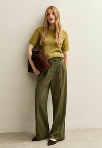 Pantalones de pierna ancha verde oliva combinados con un suéter de punto texturizado mostaza. La modelo lleva un bolso de ante marrón y calza zapatos de punta marrones.