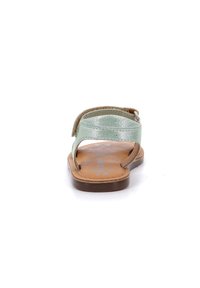 Sandalia verde menta con dos correas; suela plana de cuero marrón con patrón impreso; vista posterior que muestra un diseño sencillo y detalle de hebilla.