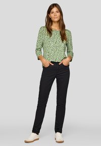 Grünes Langarmoberteil mit schwarzem Leopardenmuster, kombiniert mit schwarzen Jeans und weißen Sneakers. Das Outfit hat eine lässige Passform und eine glatte Textur.