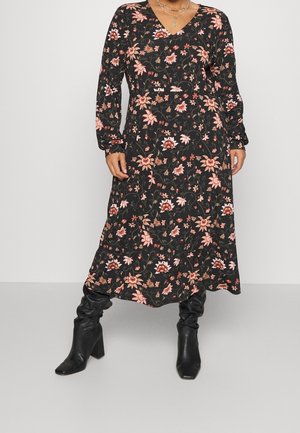 Vrouw die een zwarte midi-jurk met lange mouwen en bloemenprint met roze bloemen draagt, gecombineerd met zwarte, ruime laarzen met hoge hak en gelaagde gouden kettingen.