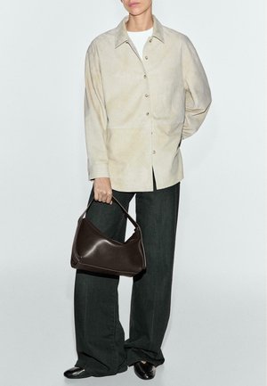 Beige ruskindsskjorte med knaplukning, kombineret med mørke bukser med vide ben. En brun lædertaske holdes i højre hånd. Sorte sko fuldender outfittet.