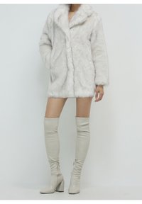Cappotto in pelliccia sintetica bianca con colletto, maniche lunghe e tasche frontali; indossato sopra stivali beige in pelle alti fino al ginocchio con tacco a blocco.